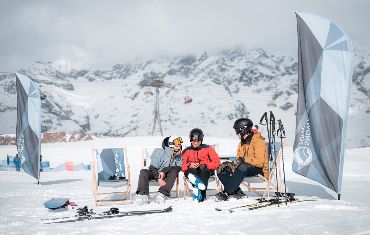 Drei Personen unterhalten sich im Snowpark auf den Liegestühlen.  | © Basic Home Production