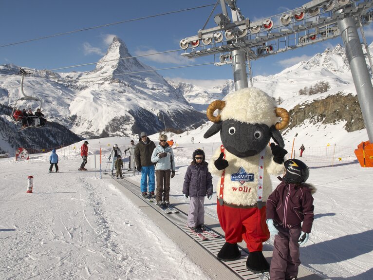 Une famille et Wolli se tiennent sur le tapis magique du parc pour débutants de Sunnegga. En arrière-plan, on aperçoit les montagnes. | © Zermatt Bergbahnen