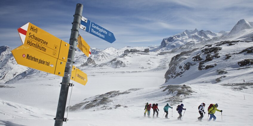 Gruppo di ciaspolatori che si fa strada nella neve, un cartello in primo piano | © Michi_Portmann