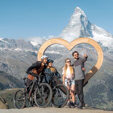 Due escursionisti e due ciclisti si scattano un selfie davanti al cuore di legno di Blauherd.  | © Basic Home Production